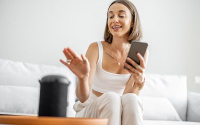 Young and cheerful woman controlling home devices with a smart speaker and phone, sitting on the couch at home